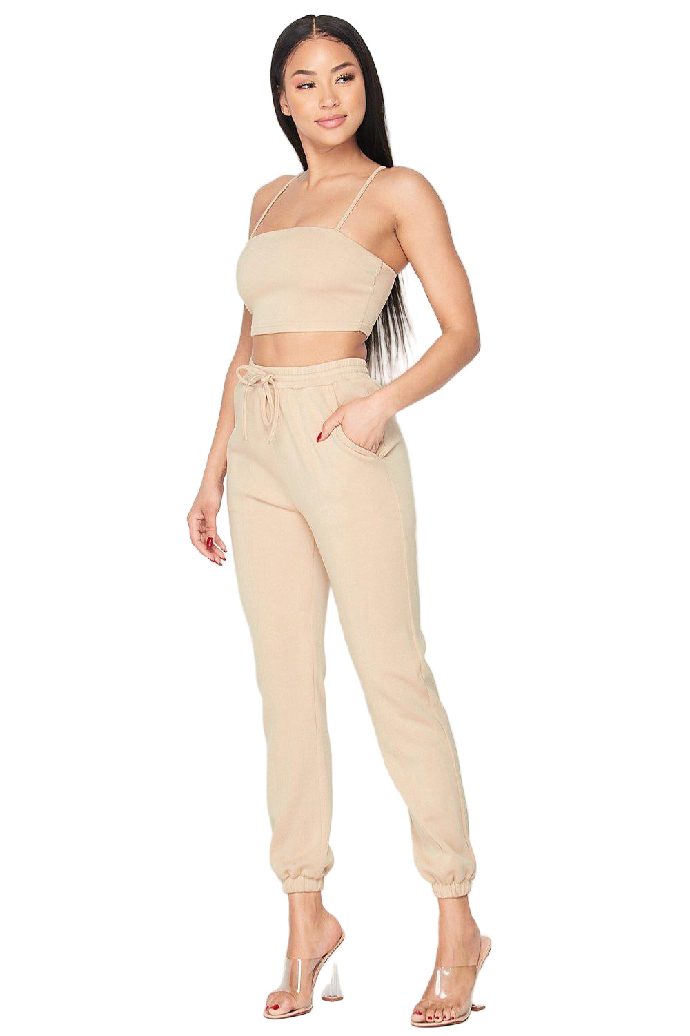 Woman wearing Keep It Cute Two-Piece Set in beige, featuring a spaghetti strap crop top and jogger pants, styled with clear heels.