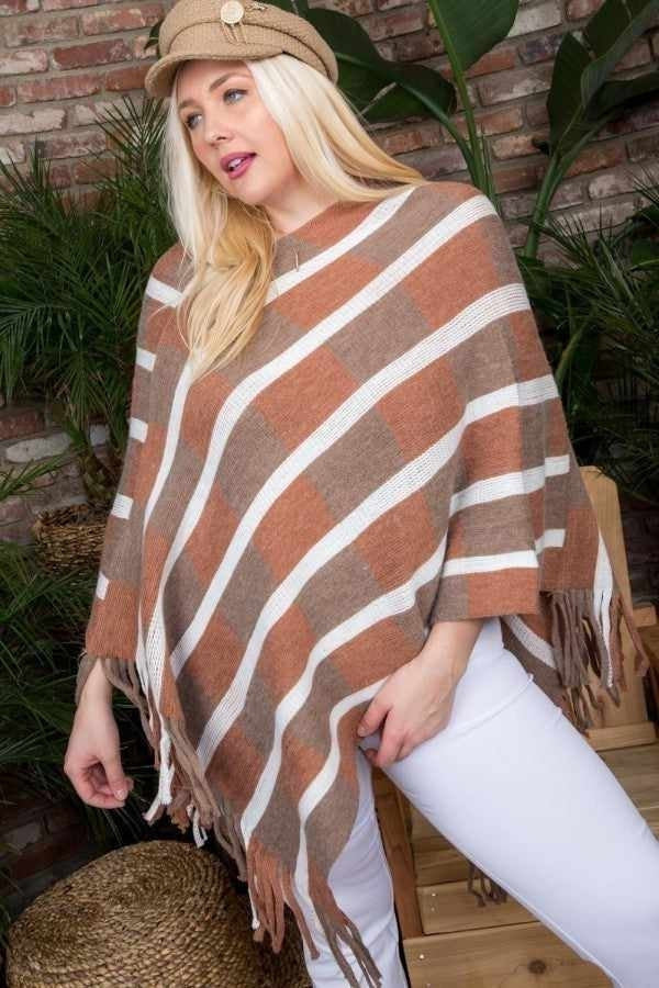 Curvy woman modeling a striped poncho-style sweater with fringe, paired with white pants and a trendy hat, set against a leafy backdrop.