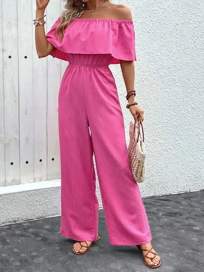 Woman wearing a stylish off-shoulder pink jumpsuit, accessorized with a woven handbag and sandals, against a minimalist backdrop.