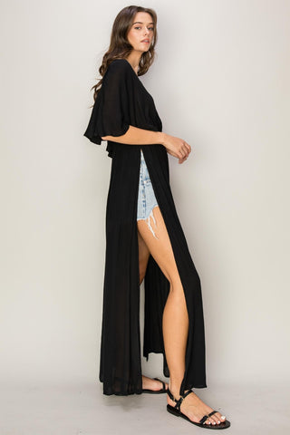 Woman wearing black tie back maxi split cover up dress, ruffled and backless, paired with denim shorts and sandals.