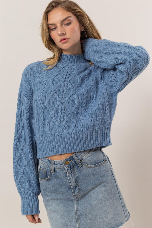 Gray blue cable-knit mock neck sweater with dropped shoulders, ribbed texture, slightly stretchy, worn with a denim skirt.