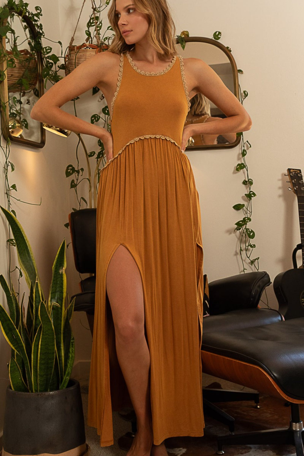 Woman wearing toffee sleeveless maxi dress with front slit, surrounded by plants and a chair.