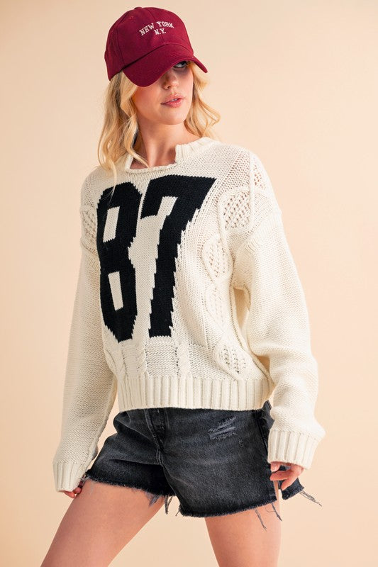 Woman wearing a white cable knit sweater with a black number 87 design and a red cap, paired with black denim shorts.