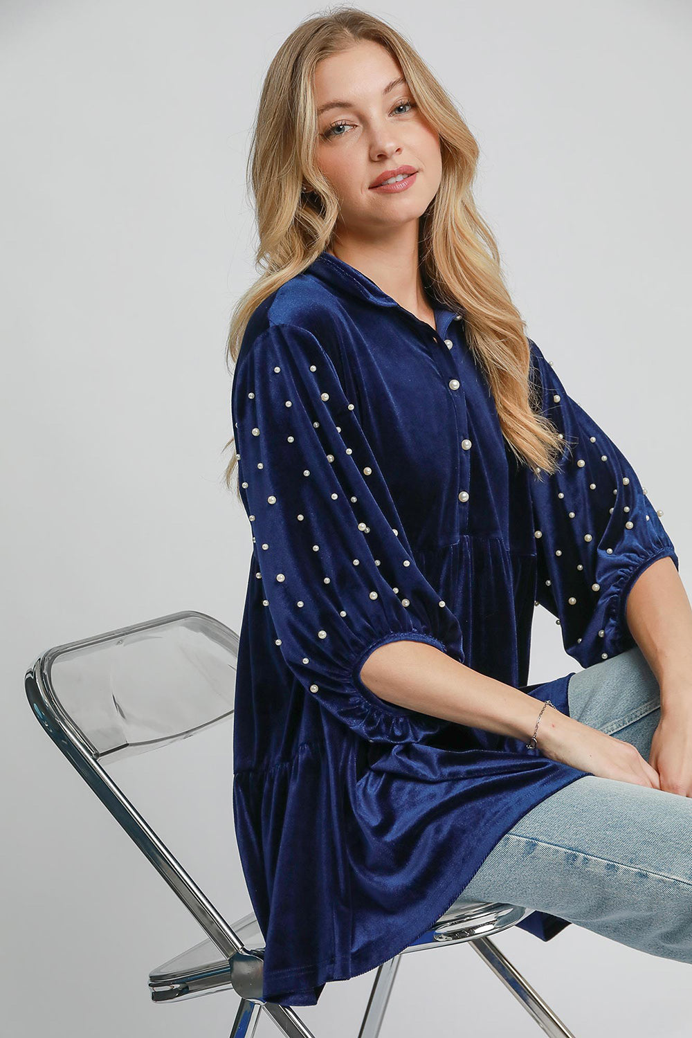 Woman wearing navy velvet babydoll blouse with pearl details, sitting on a chair.