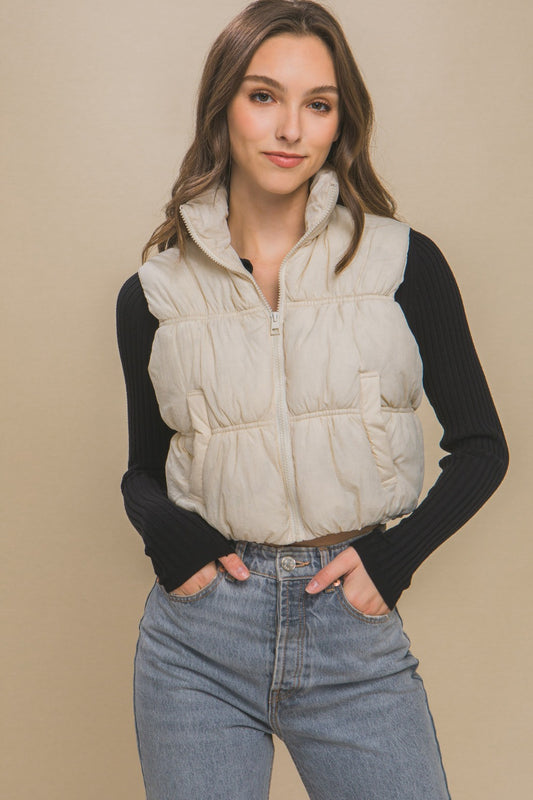 Woman wearing cream zip-up turtleneck cropped vest coat over black long-sleeve top and jeans.