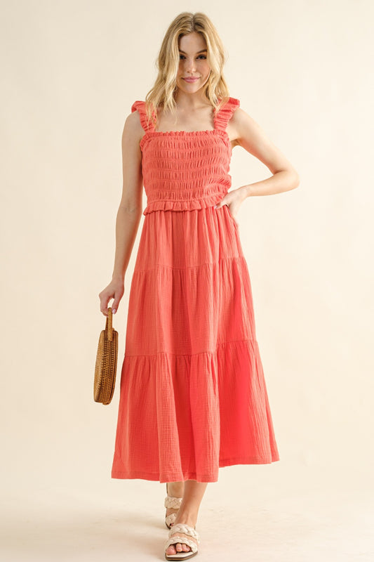 Woman wearing coral smocked ruffled tiered dress made of 100% cotton, holding a wicker bag, stylish summer fashion.