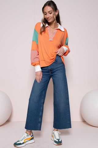 Woman wearing coral color block top with striped panel, paired with wide-leg jeans and sneakers, standing against a light backdrop.