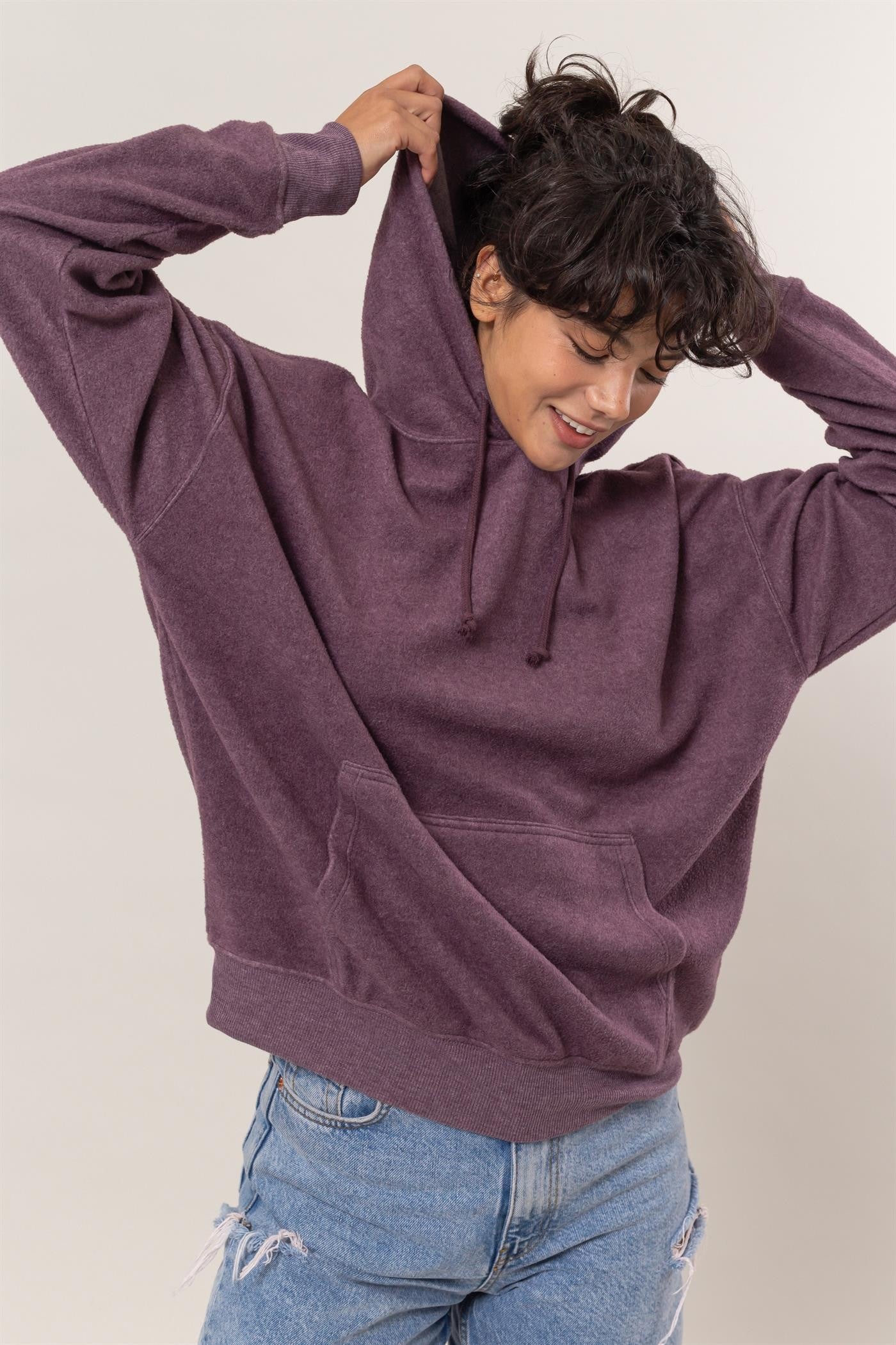 Woman wearing a dark plum long sleeve hoodie with kangaroo pocket, slightly stretchy, made of polyester and rayon, pulling up hood.