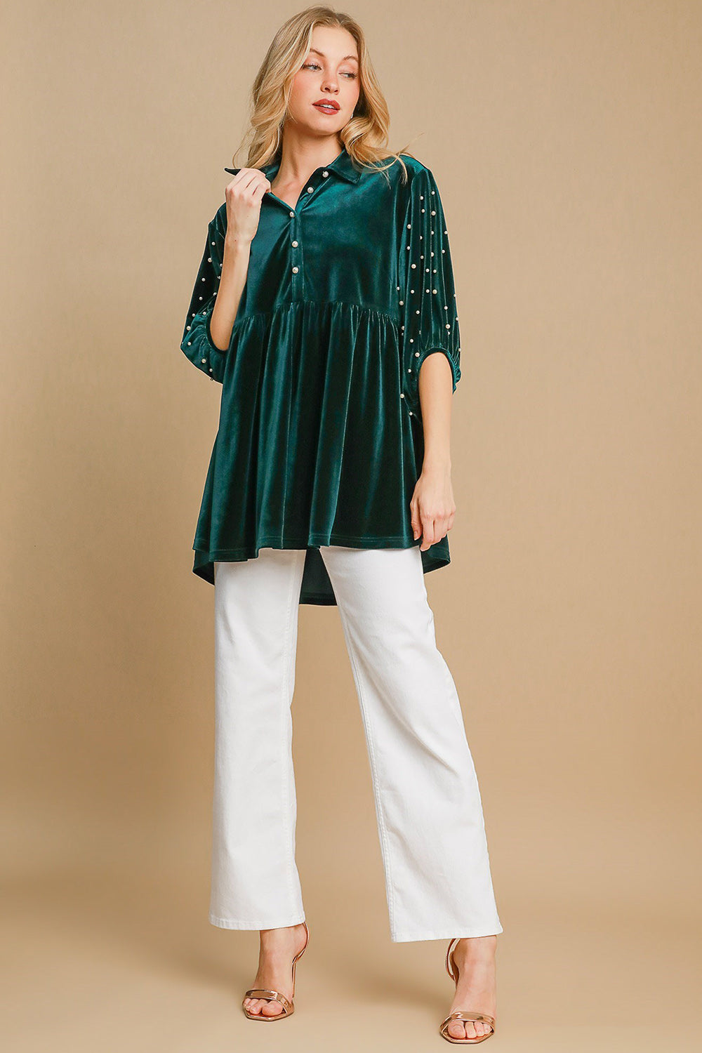 Woman wearing hunter green velvet babydoll blouse with buttoned, tiered design and white pants, standing on beige background.