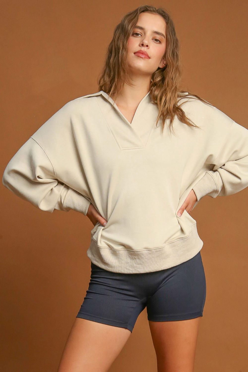 Woman wearing a Johnny Collar Dropped Shoulder Sweatshirt with navy shorts against a brown background.