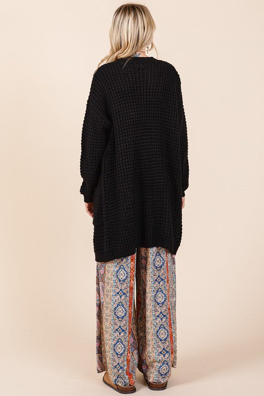 Woman wearing a black open front long sleeve longline cardigan with waffle-knit texture, paired with patterned pants.