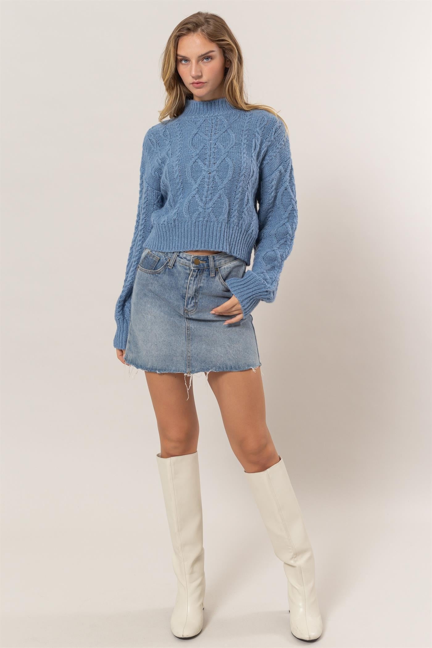 Woman in gray blue cable-knit mock neck sweater with denim skirt and white boots.