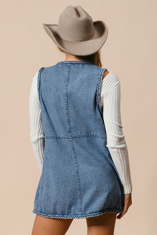 Woman wearing a braided trim open front denim vest with pockets and a beige hat, showcasing the back view of the clothing piece.