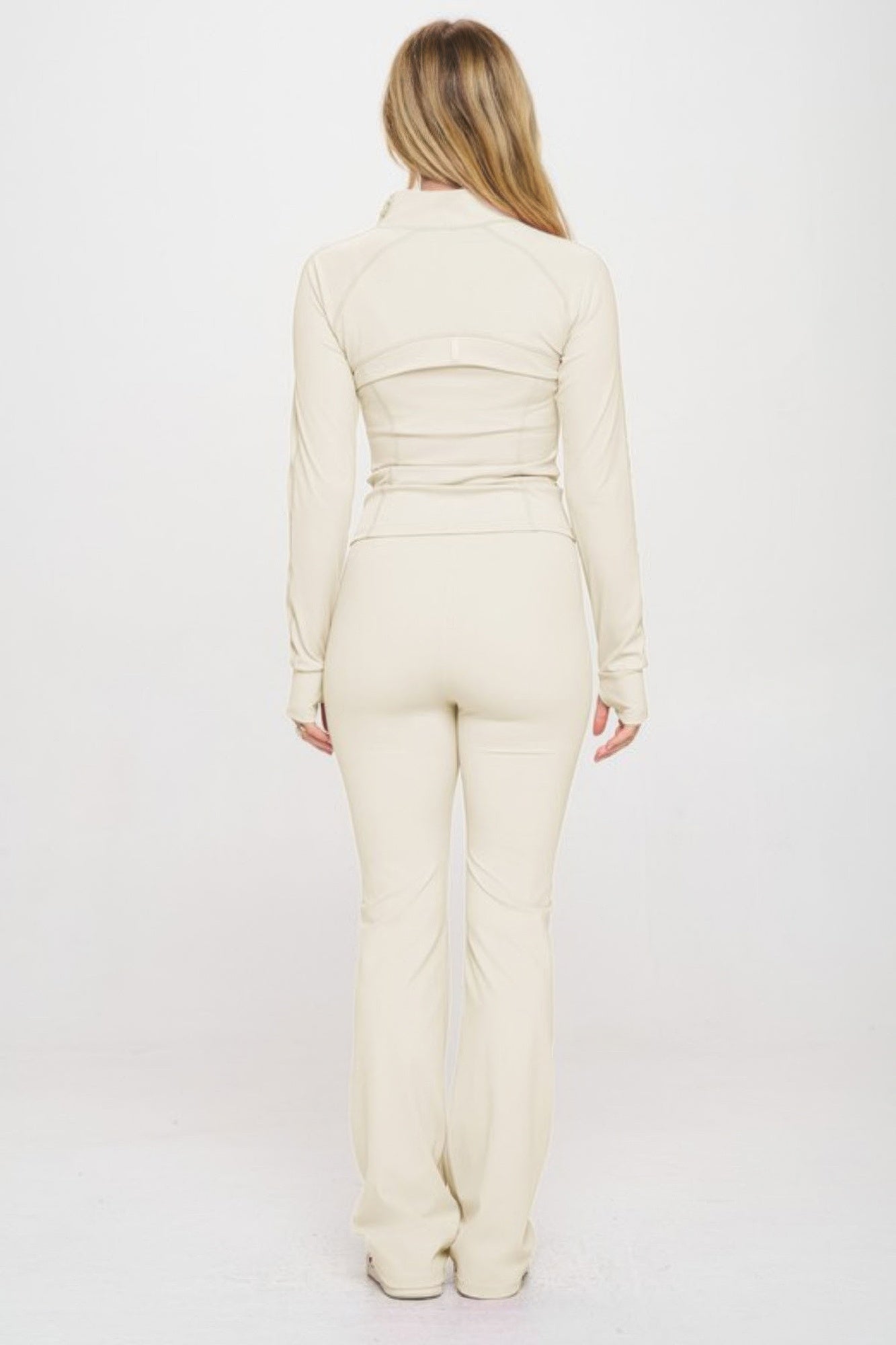 Woman wearing beige fitted yoga jacket with thumbholes and matching flare-bottom jumpsuit from the back view.