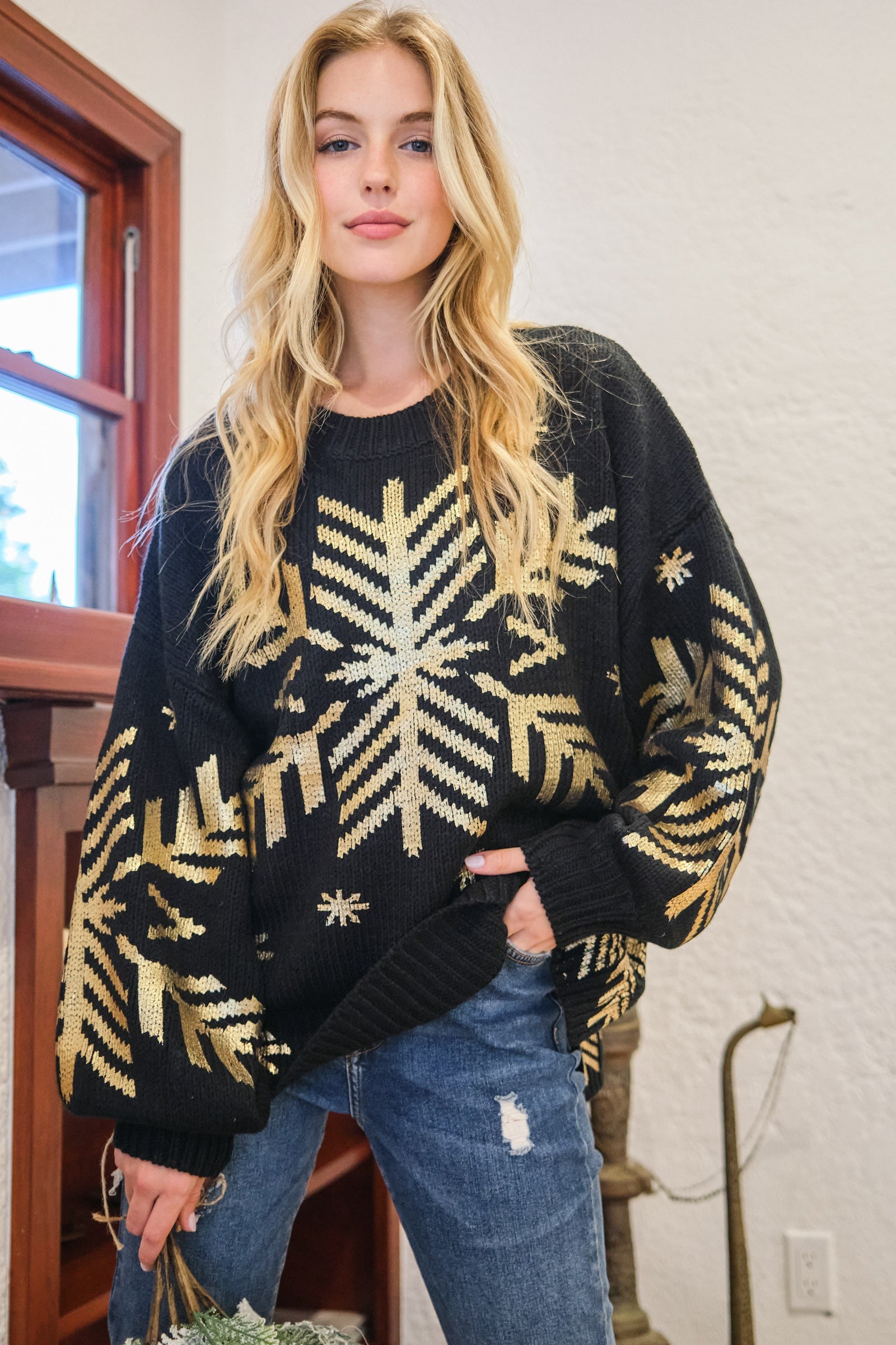 Woman wearing a black foil snowflake round neck sweater with ribbed hems, slightly stretchy fabric, paired with jeans indoors.