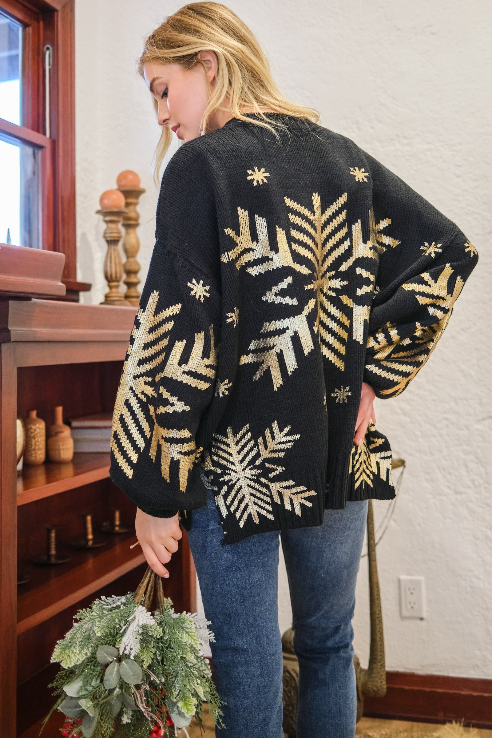 Woman wearing black sweater with large gold foil snowflake design, standing near wooden shelf holding greenery in a cozy room.