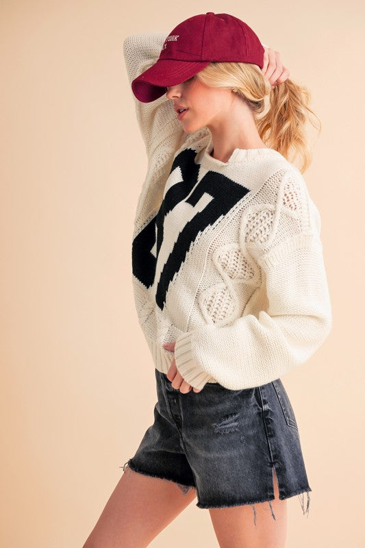 Woman wearing a white cable knit drop shoulder sweater with black contrasting number, paired with denim shorts and a red cap.