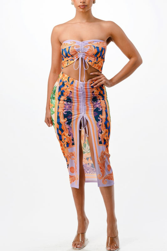 Woman in strapless bandeau top and midi skirt with colorful baroque print in orange, blue, and purple. Both pieces feature front tie details and...