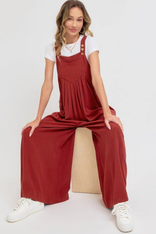 Woman wearing a chic adjustable strap overall wide leg jumpsuit in red with a relaxed fit, sitting on a wooden block.