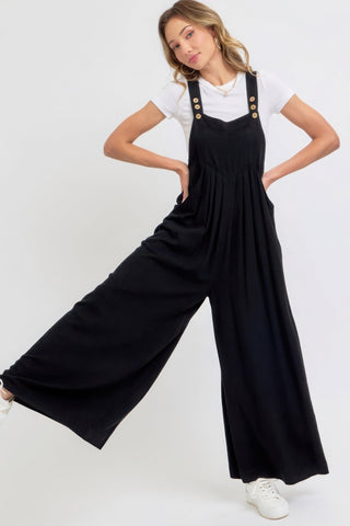 Woman wearing a black adjustable strap overall wide leg jumpsuit with a white shirt and sneakers, featuring a modern slouchy silhouette.