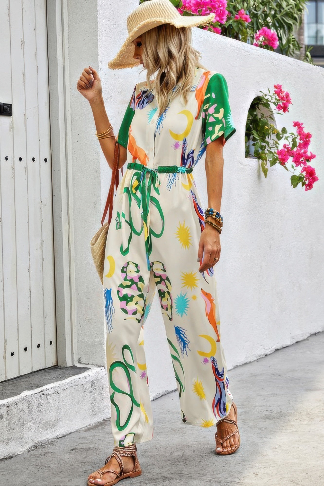 Woman in colorful printed maxi dress with green belt and straw hat walking past white building with pink bougainvillea flowers. She's wearing...