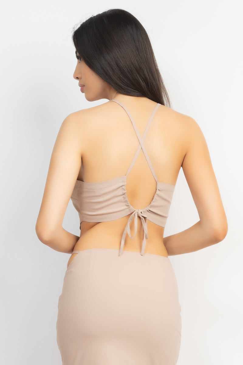 Back view of a woman wearing a light taupe asymmetrical cross back top with drawstring closure and matching high-waist skirt.