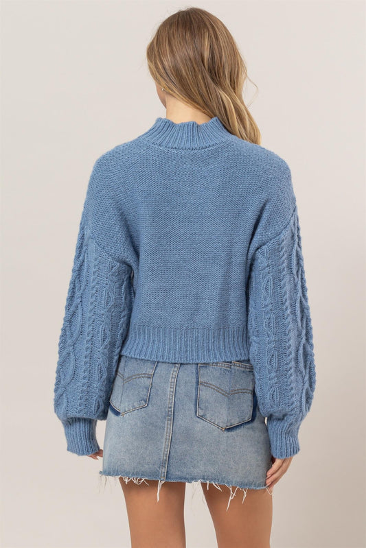 Woman wearing gray blue cable-knit mock neck sweater with dropped shoulders and a denim skirt, back view