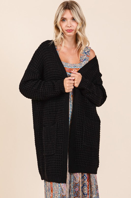 Black waffle-knit longline cardigan with pockets and ribbed texture, worn over a patterned dress.