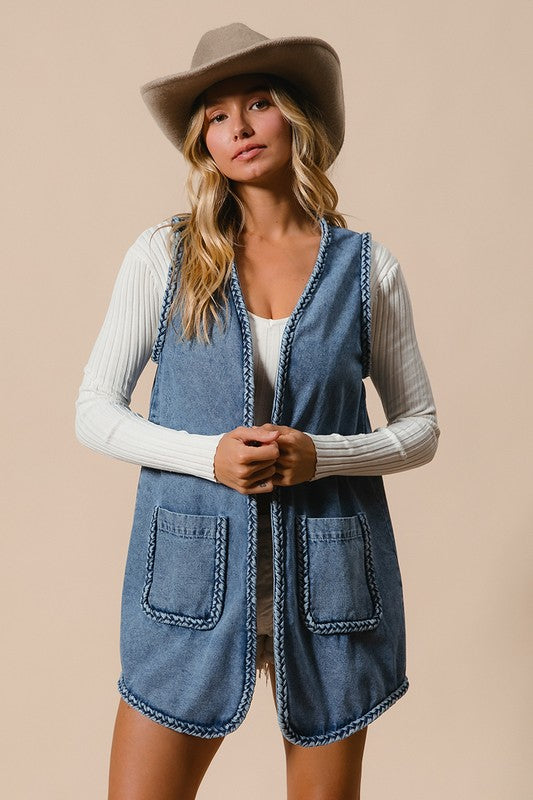 Woman wearing a braided trim open front denim vest with pockets, styled over a white ribbed top, and accessorized with a cowboy hat.