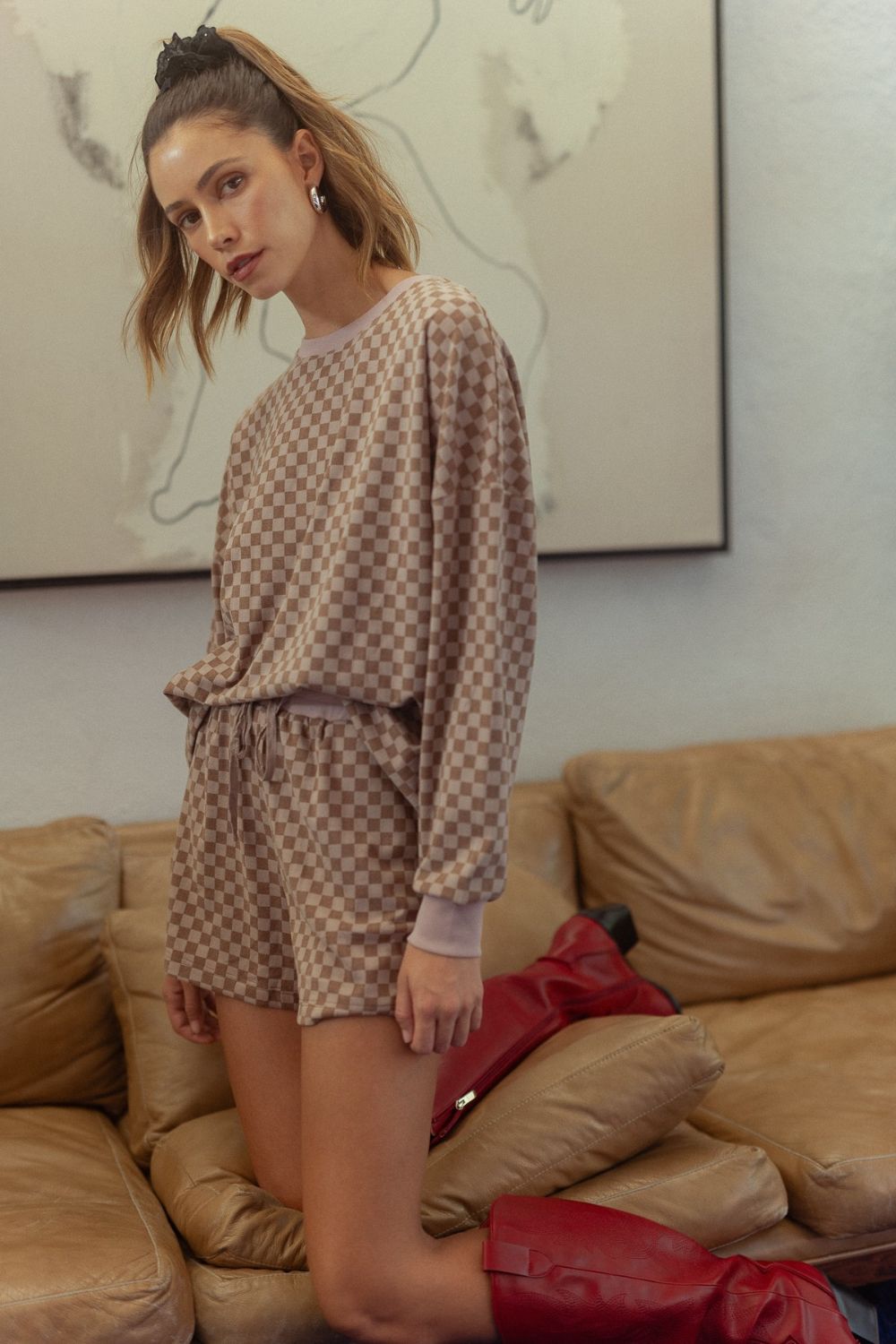 Woman wearing mocha checkered round neck top and drawstring shorts set with red boots, standing on a beige couch.