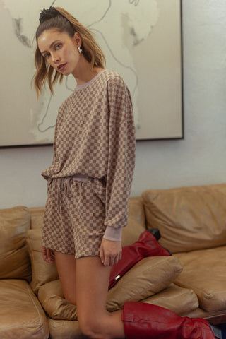 Woman wearing mocha checkered round neck top and drawstring shorts set with red boots, standing on a beige couch.