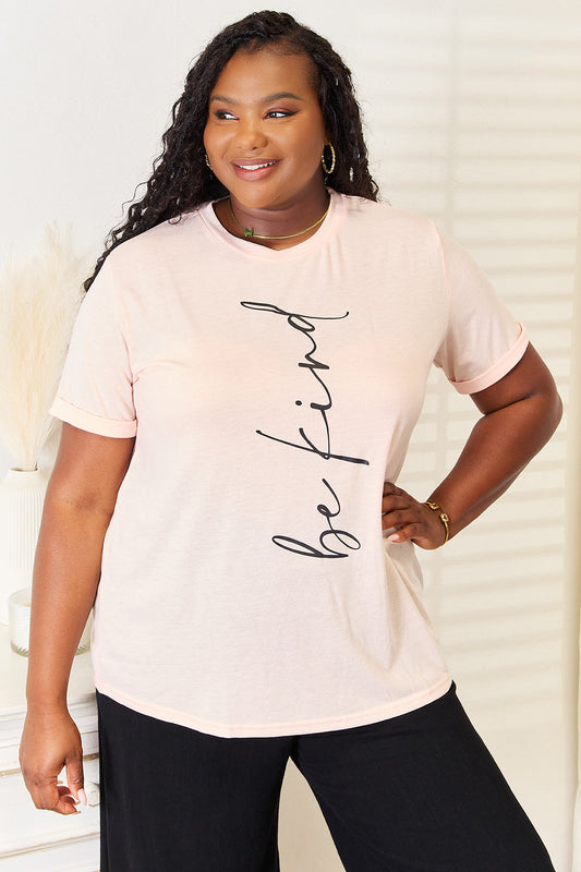 Woman wearing a BE KIND graphic round neck T-shirt in soft pastel, paired with casual black pants.