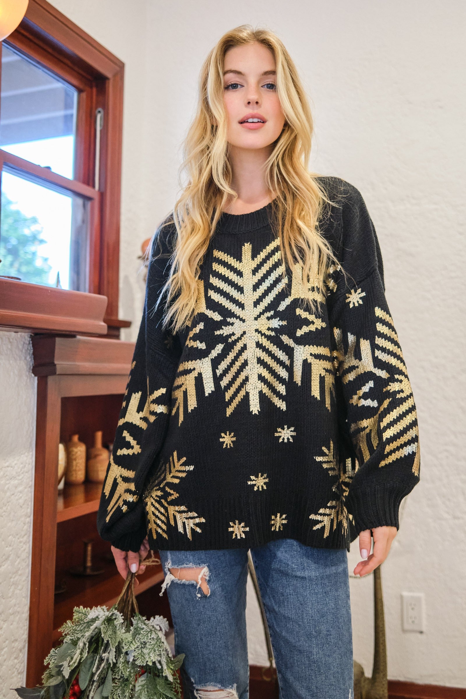 Woman wearing a foil snowflake round neck sweater in black, paired with distressed jeans, in a cozy indoor setting.