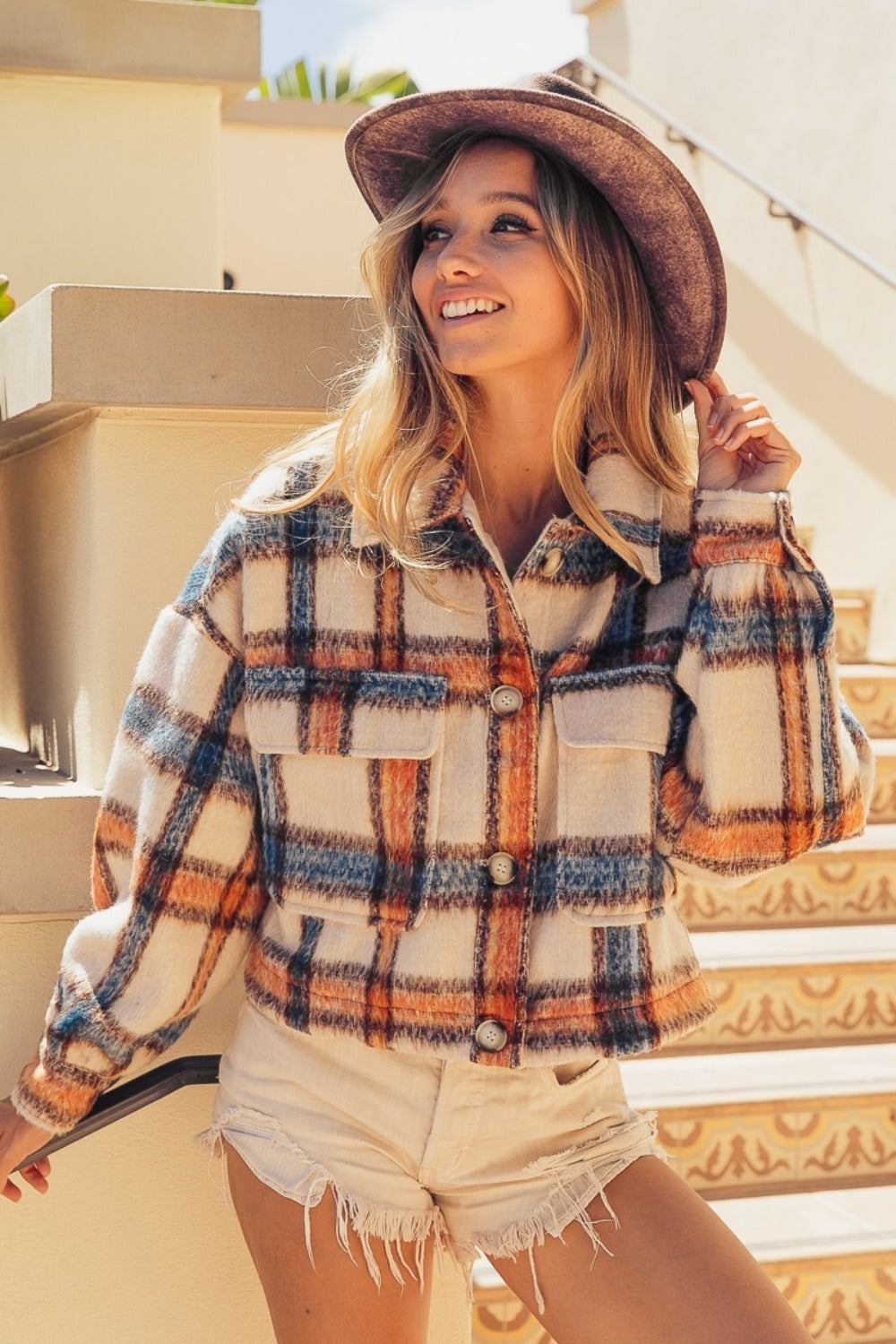 Woman wearing an ivory multi-color brushed plaid crop jacket with pockets, standing on stairs, paired with a hat and white shorts.