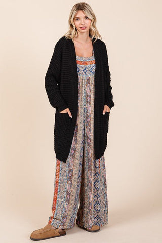 Black open front long sleeve longline cardigan with pockets and ribbed waffle-knit, worn over patterned jumpsuit.