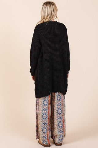 Black ribbed waffle-knit longline cardigan with pockets and long sleeves, shown from the back on a person in patterned pants.