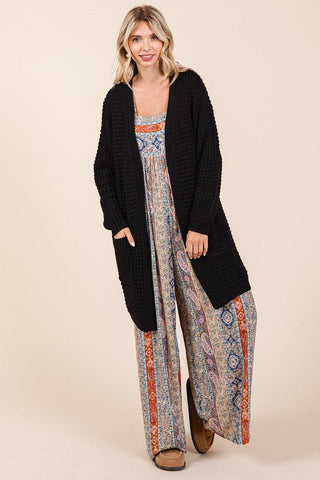 Woman wearing black open front longline cardigan over patterned dress with long sleeves and pocketed design.
