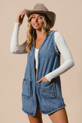 Woman wearing braided trim open front denim vest with pockets and a beige hat.