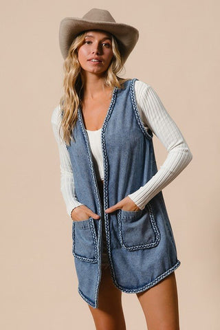 Woman wearing braided trim open front denim vest with pockets over a white top, styled with a beige hat.