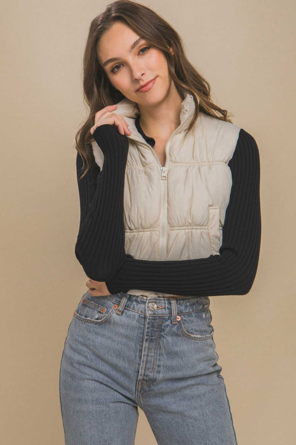 Woman wearing cream zip-up turtleneck cropped vest coat, black long sleeve top, and jeans.