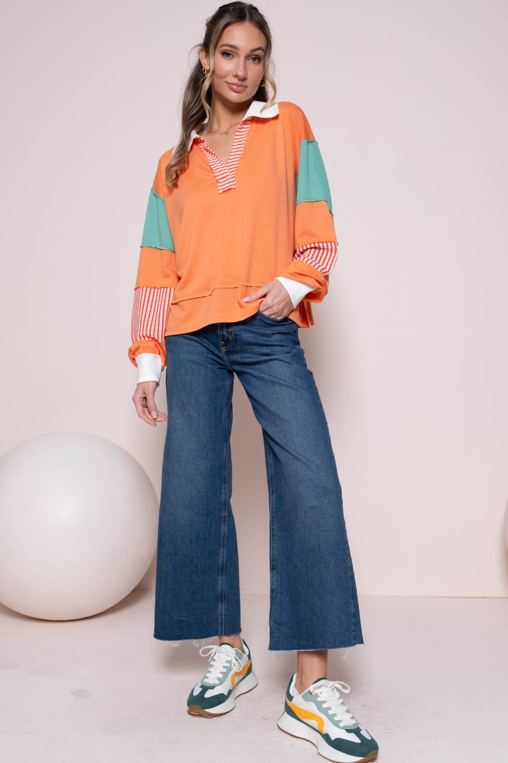 Woman wearing coral color block top with striped panel and wide-leg jeans standing in a minimalistic room.