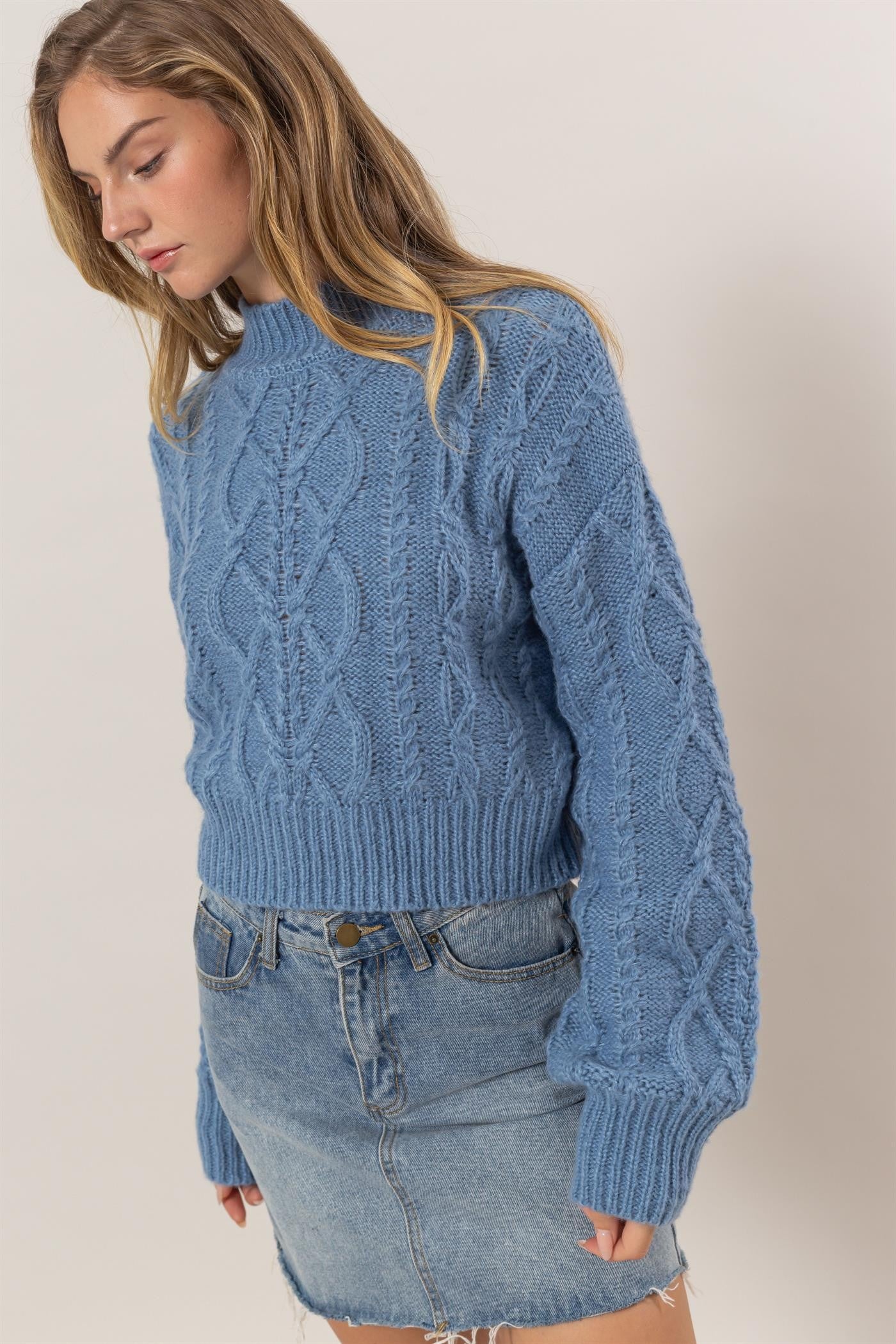 Woman wearing a gray blue cable-knit mock neck sweater with ribbed edges, paired with a denim skirt.