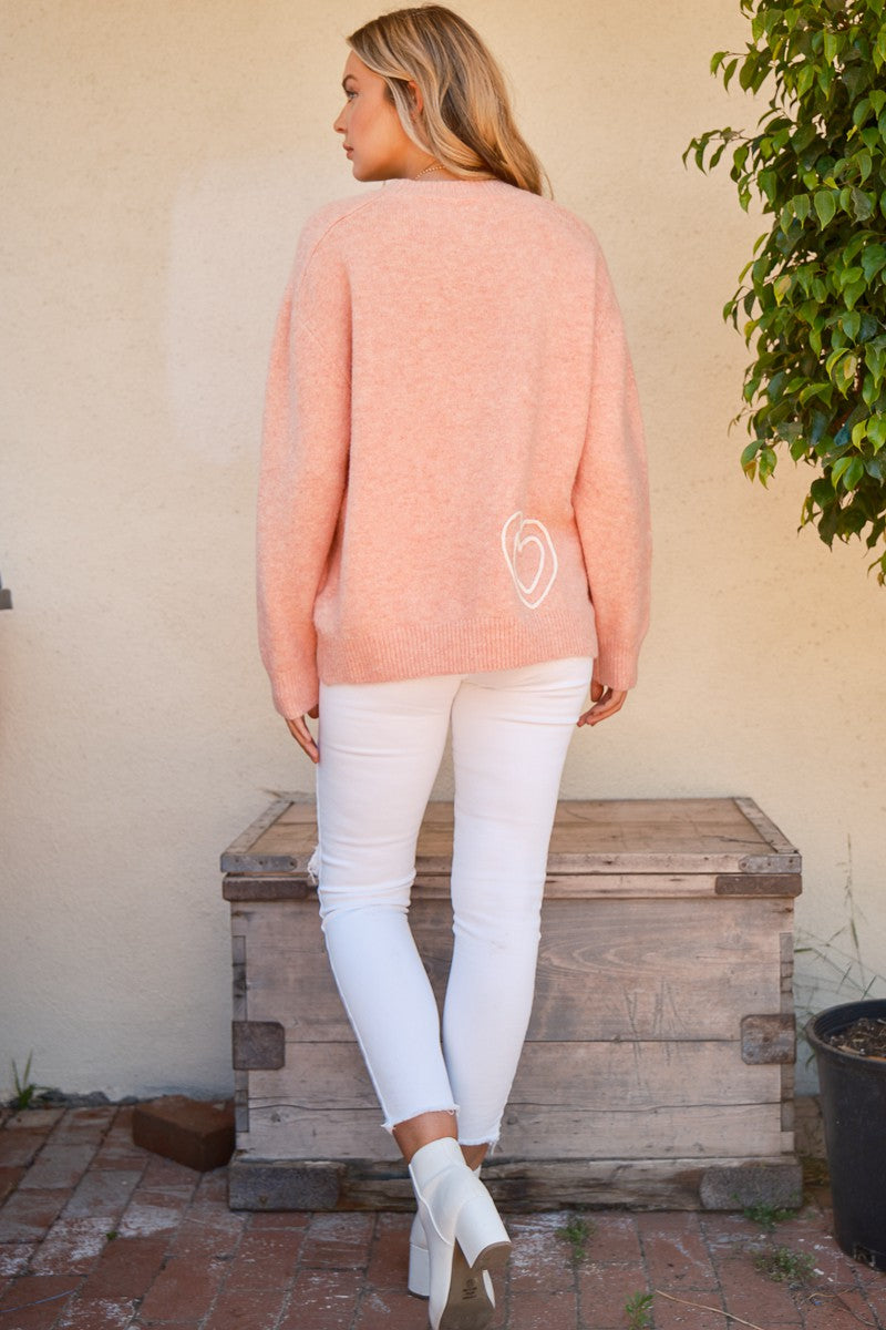 Woman wearing pink round neck sweater with heart design, paired with white pants and boots, standing outdoors.