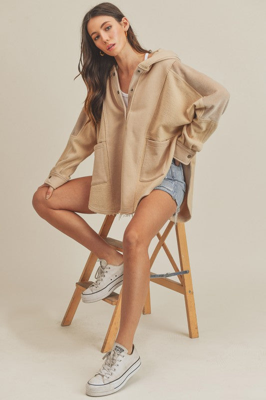 Woman wearing an Oat Slit Half Snap Oversize Hoodie with ribbed raw hem and pockets, seated on a wooden stool.