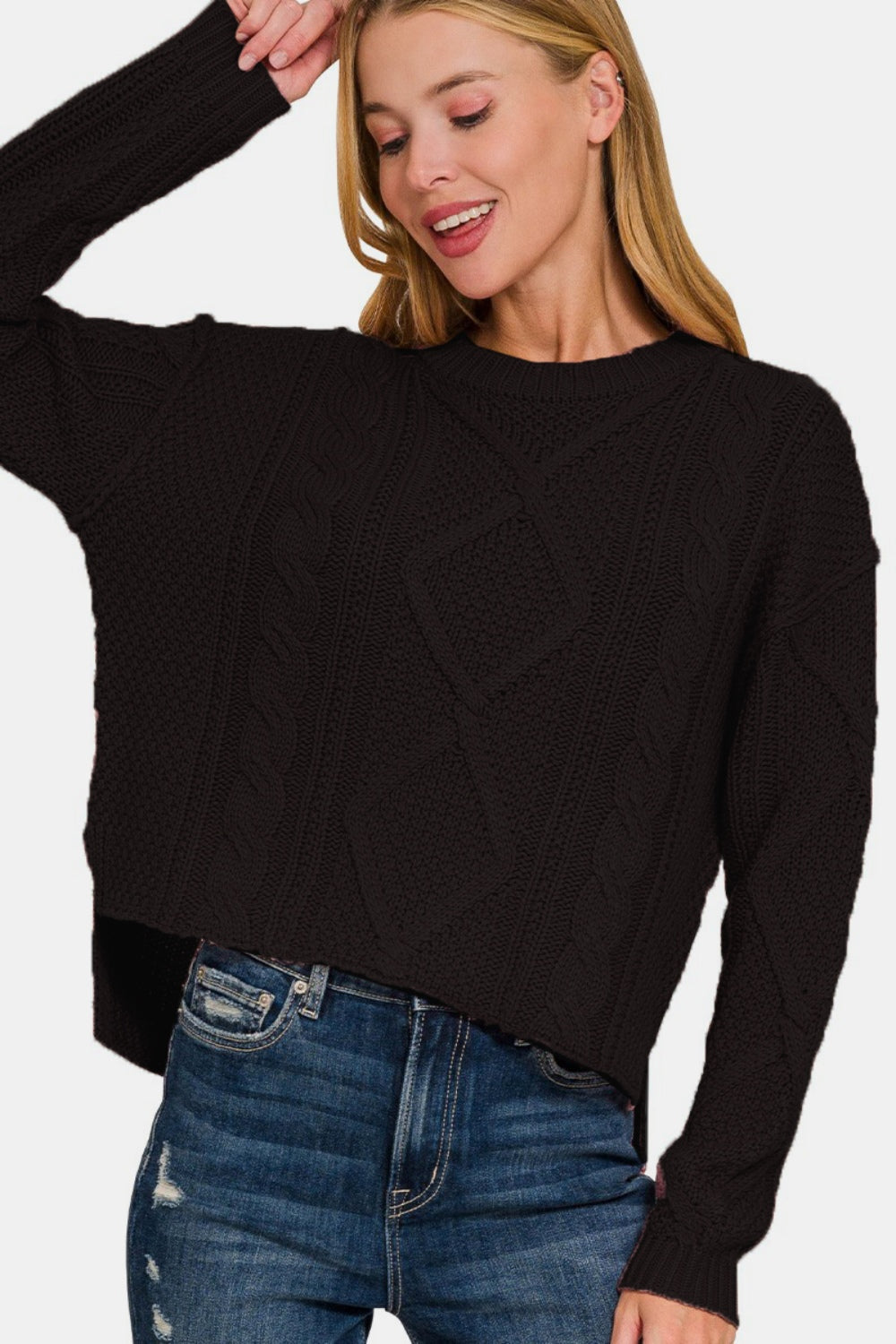 Woman wearing cropped high-low cable sweater with side slits, featuring an exposed seam detail and cozy knit design.