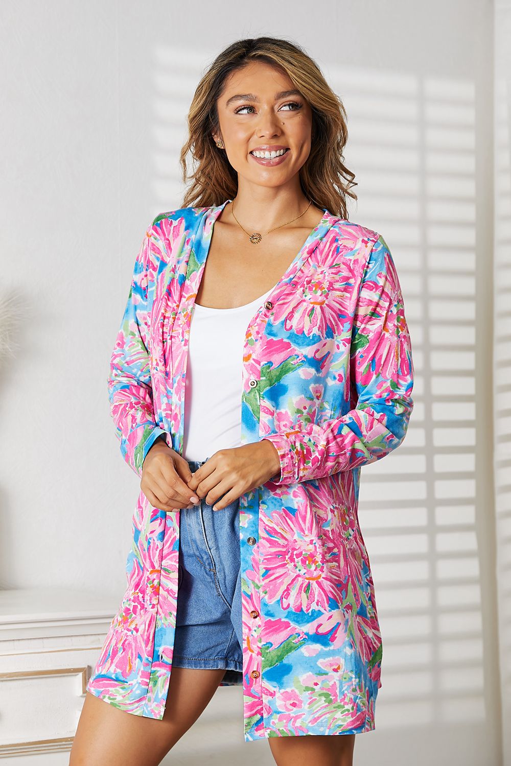 Woman wearing a vibrant floral open front cardigan with long sleeves over a white top and denim shorts, standing indoors.