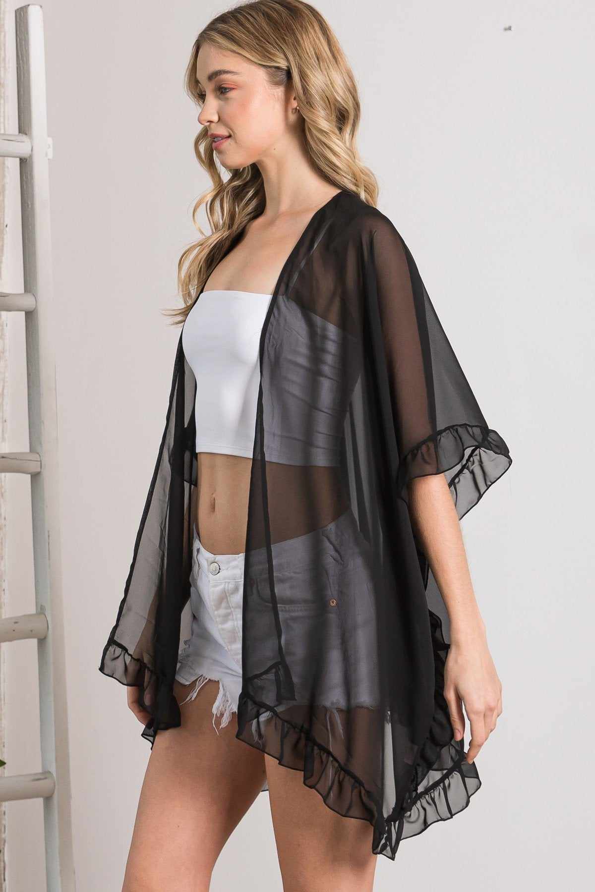 Woman wearing a black lightweight sheer shawl cardigan with ruffle edges, styled over a white top and denim shorts.