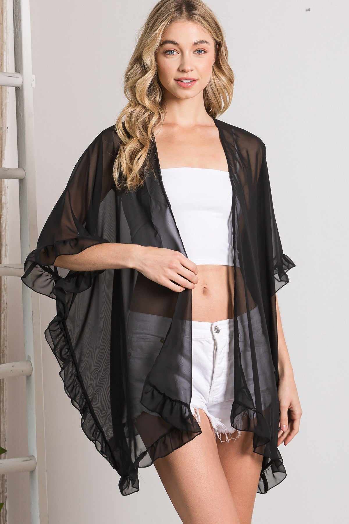 Woman wearing a black lightweight sheer shawl cardigan with ruffle edges, styled with a white top and shorts for an elegant look.