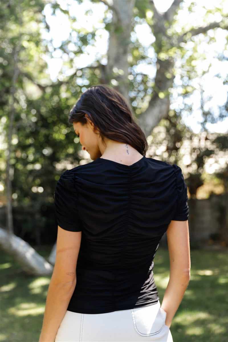 Woman wearing a black mesh ruched cut-out short sleeve top, standing outdoors with trees in the background.