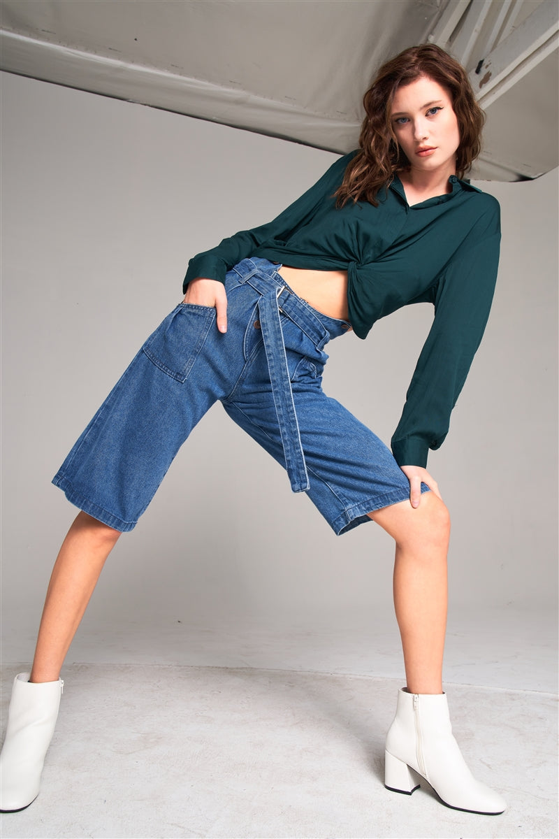 Woman wearing mid blue denim midi flare jeans with high waist, buckle belt, styled with green blouse and white boots for a vintage look.